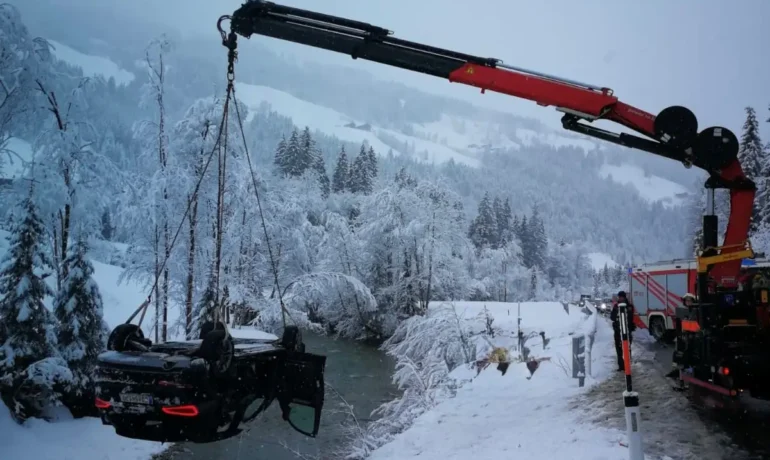 Kran hebt im verschneiten Winter eine verunglückte, auf dem Dach liegende schwarze Auto aus einem Flussbett bei Haitzmann