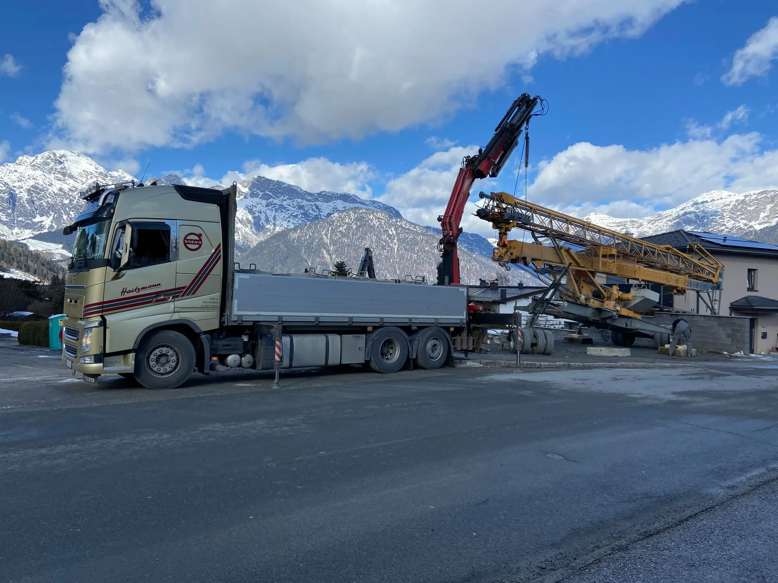 LKW mit Kran hebt Bauteil vor Haus in Kleinarl vor Bergkulisse mit Schnee und blauem Himmel