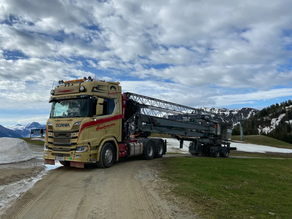 Haitzmann Transporte Schwerlast-LKW mit Kran auf Bergstraße vor schneebedeckten Alpen und bewölktem Himmel in Kleinarl