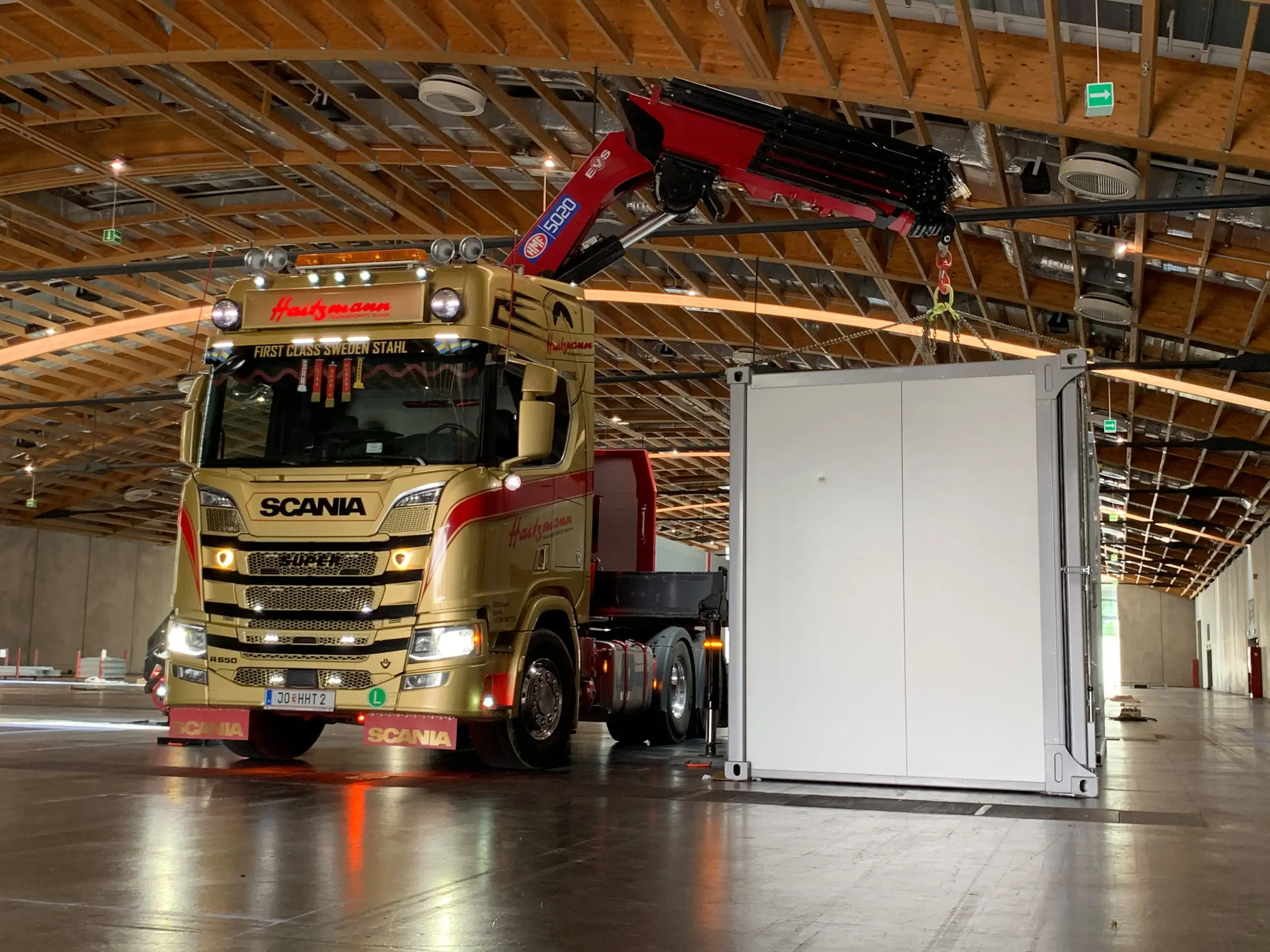 Goldfarbener Scania LKW mit Kran hebt weißen Container in großer Halle bei Haitzmann Transporte in Kleinarl.