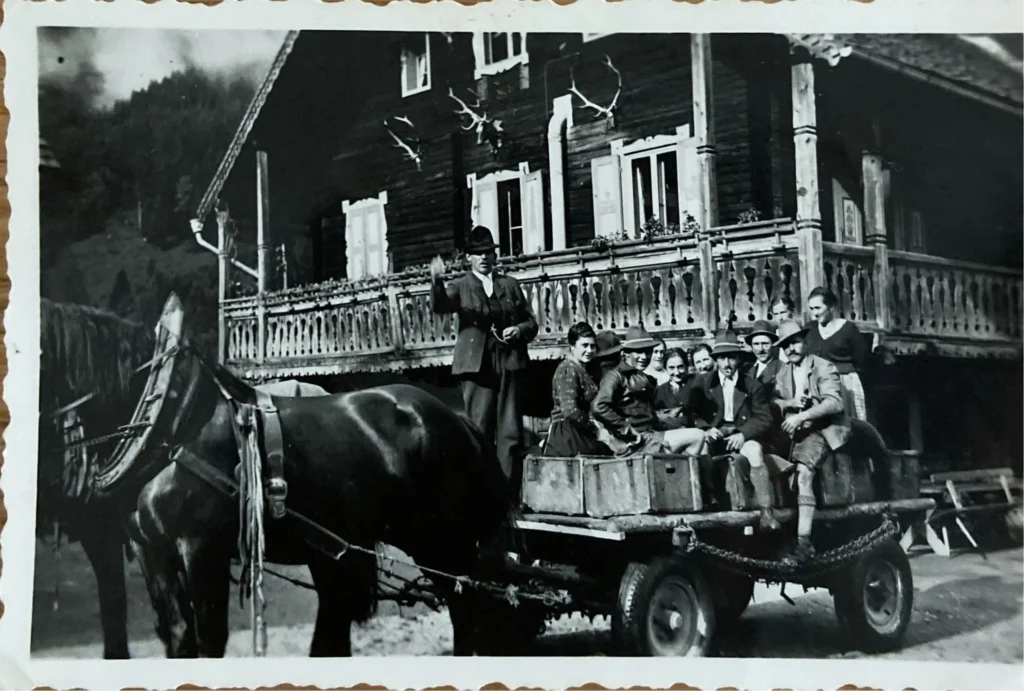 Schwarzweißfoto von neun Personen auf einem Pferdewagen vor einem traditionellen Holzhaus mit Geweihen an der Fassade in