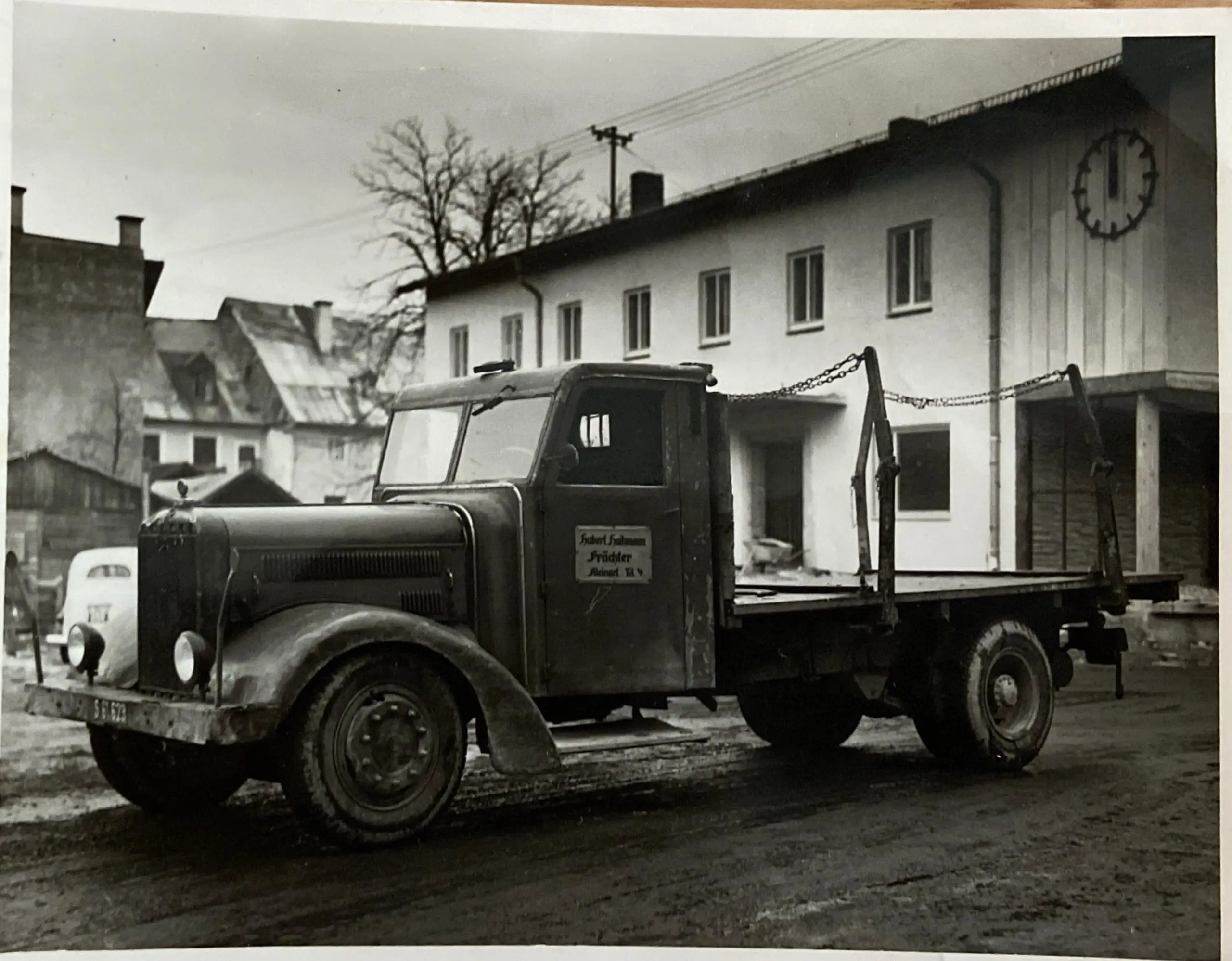 Schwarz-weiß Foto eines alten Lastwagens mit Flachbett vor einem Gebäude mit Uhr, vermutlich 1948 in Kleinarl, Transporte