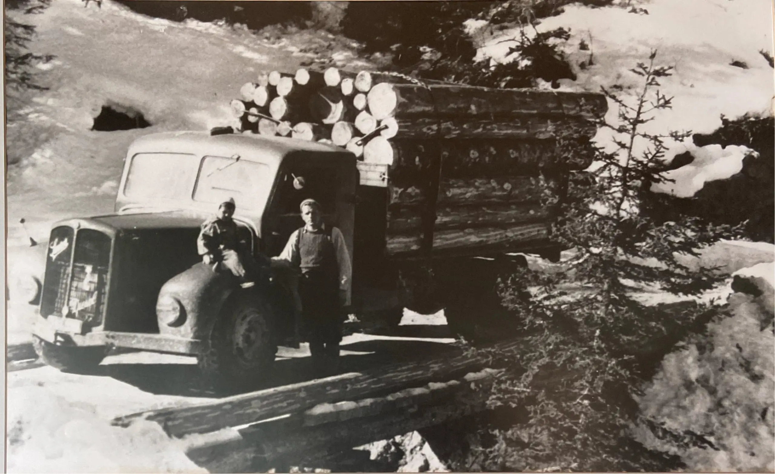 Schwarzweißfoto von zwei Männern mit einem alten Holztransporter beladen mit Baumstämmen in verschneiter Berglandschaft bei