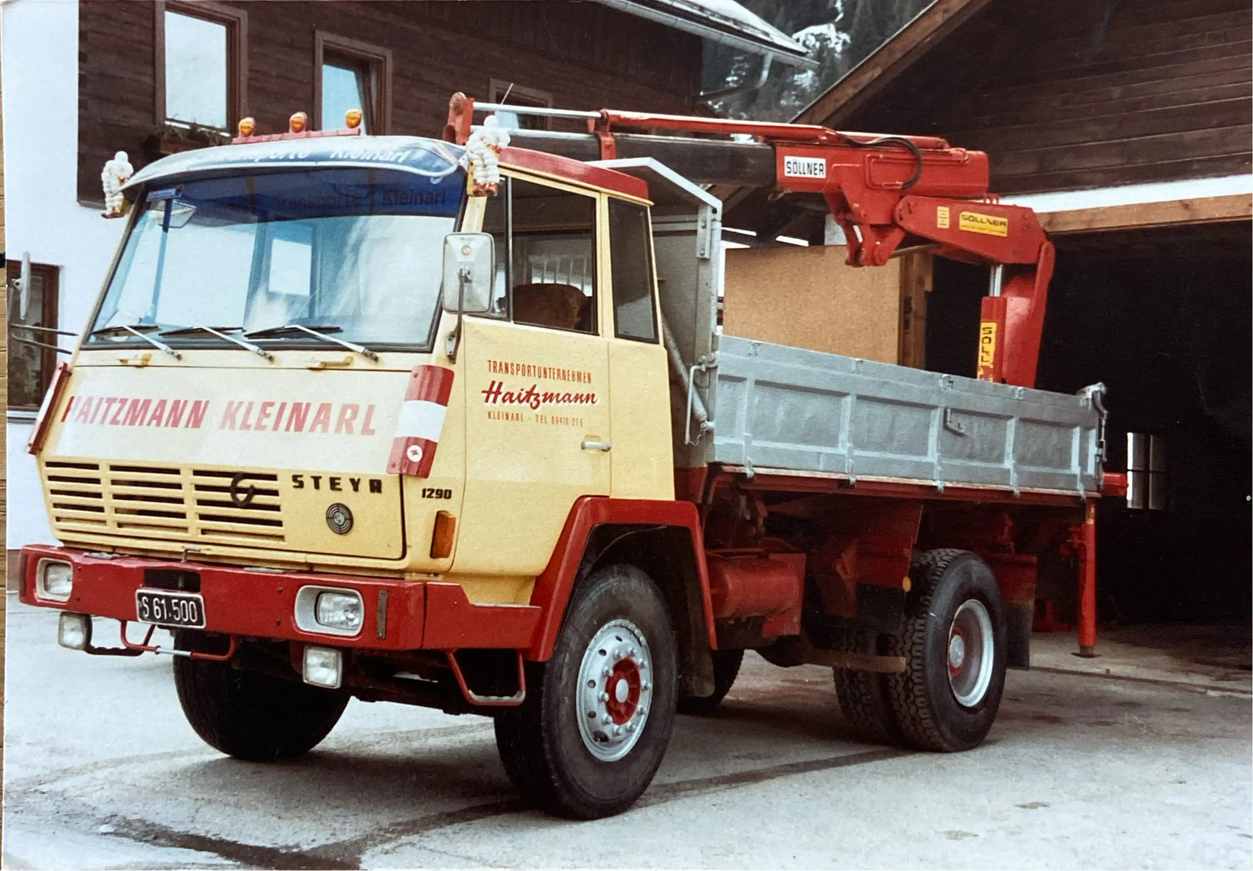 Gelb-roter Kran-LKW der Firma Haitzmann Transporte Kleinarl vor einem Holzgebäude, Modell Steyr 1290, Baujahr 1976.
