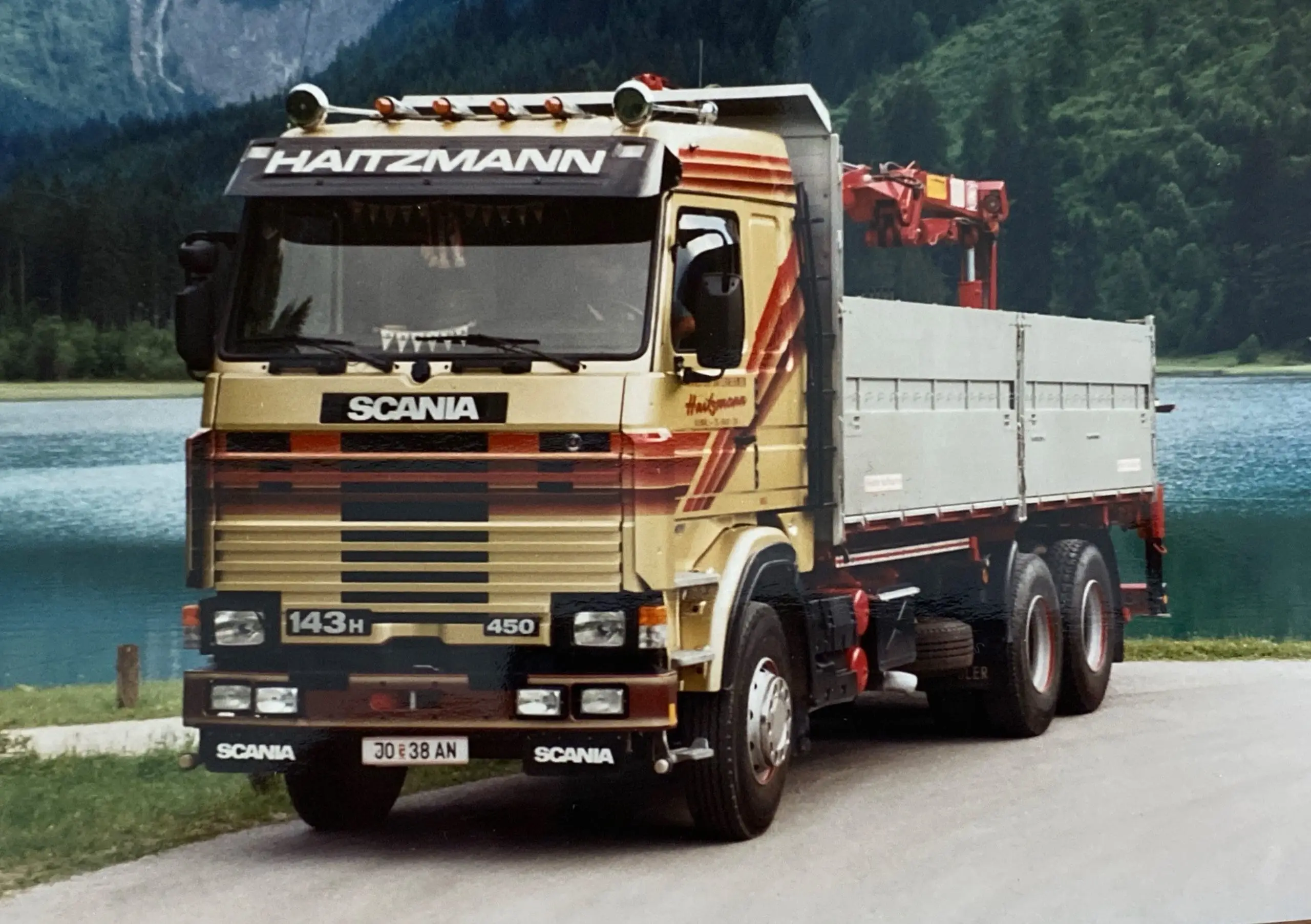 LKW der Marke Scania mit Aufschrift Haitzmann steht an einem See vor bewaldeten Bergen, Bau- und Kranarbeiten sichtbar.