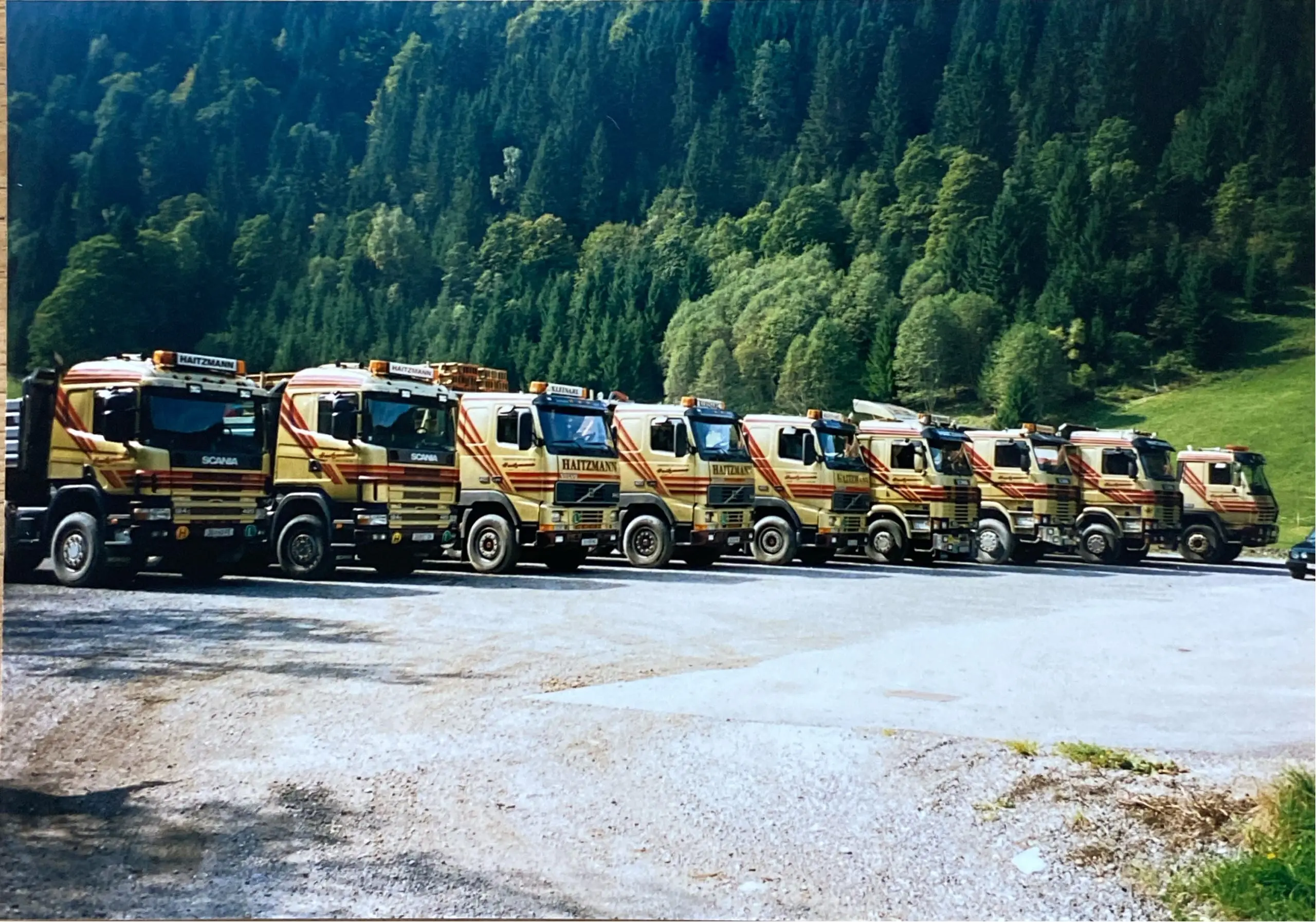 Reihe von neun gelb-roten Haitzmann Transporte LKWs vor bewaldetem Berghang auf Schotterparkplatz in Kleinarl.