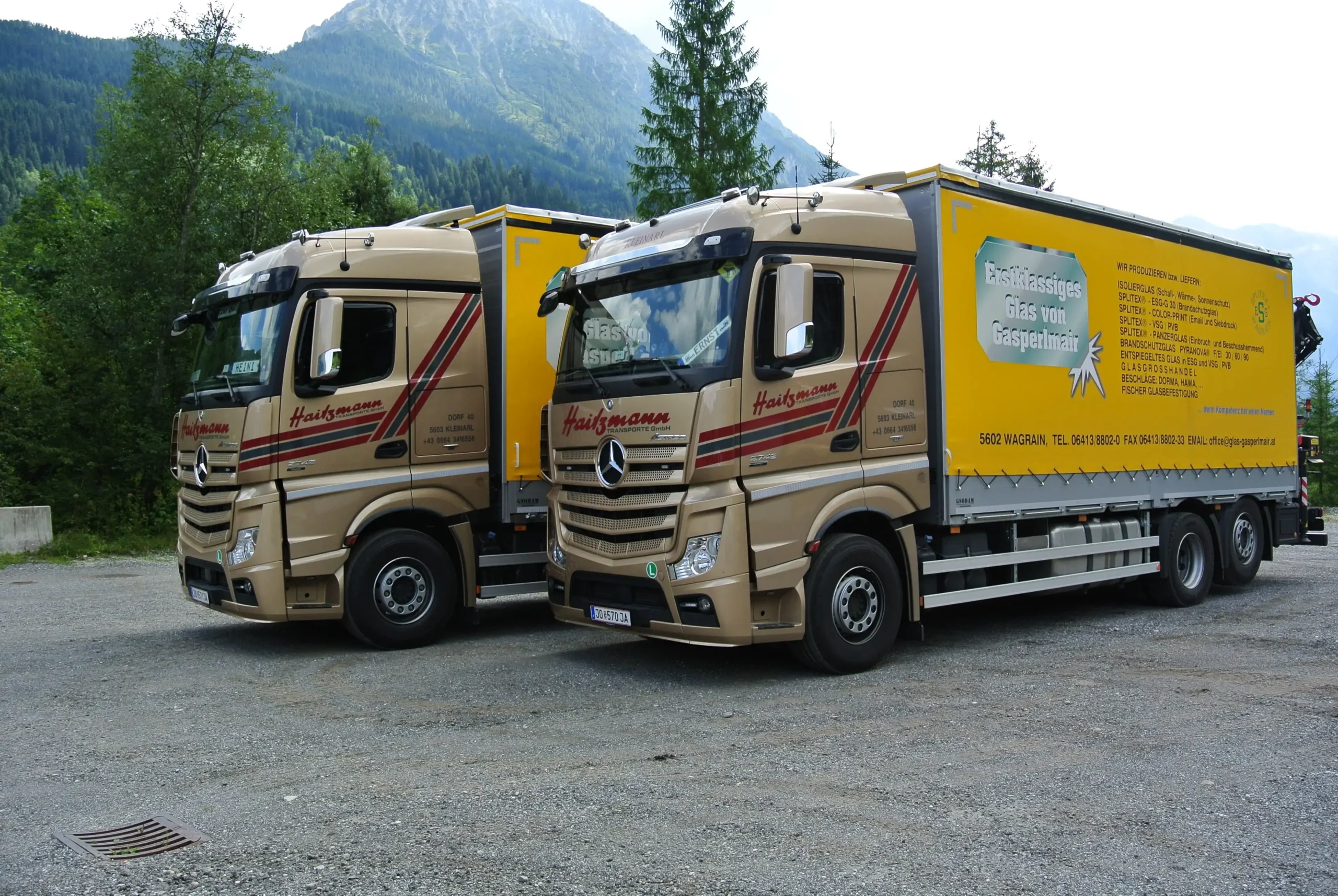 Zwei beige Haitzmann Transporte LKWs mit gelben Anhängern stehen auf einem Parkplatz vor bewaldeten Bergen in Kleinarl.