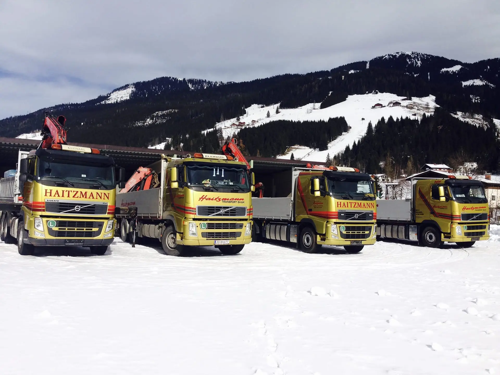 Vier gelbe Haitzmann Transporte LKWs mit Kranaufbau stehen auf schneebedecktem Gelände vor verschneiten Bergen in Kleinarl.